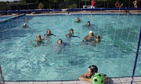 People swim in a pool playing water polo, surrounded by a mesh net, under a sunny sky at Oasi