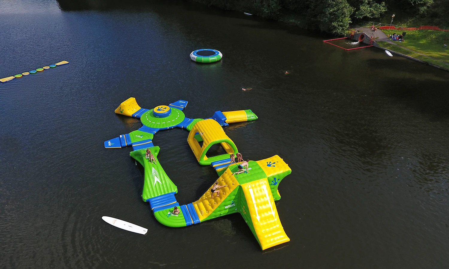 Inflatable water park with people climbing and sliding, floating on a calm lake near a wooded shore at Natterer See