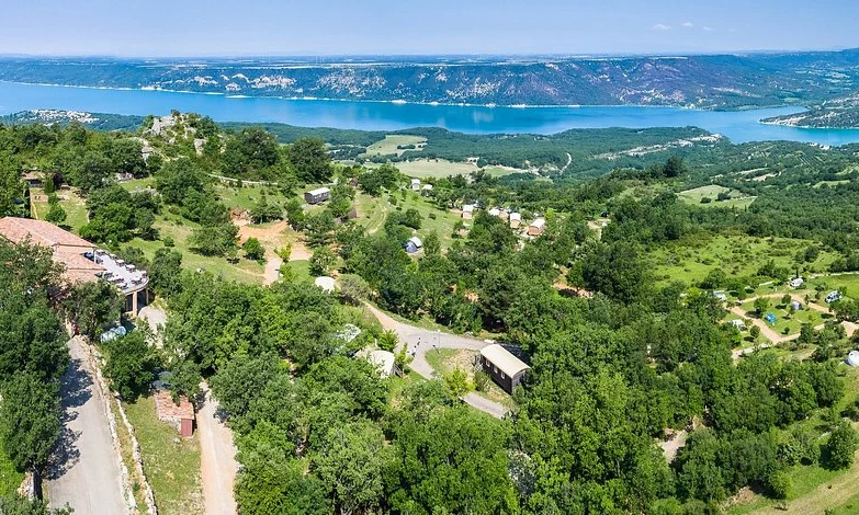 Houses and trees atop a hilly area with a winding road, overlooking a serene blue lake and distant mountains at de l'Aigle