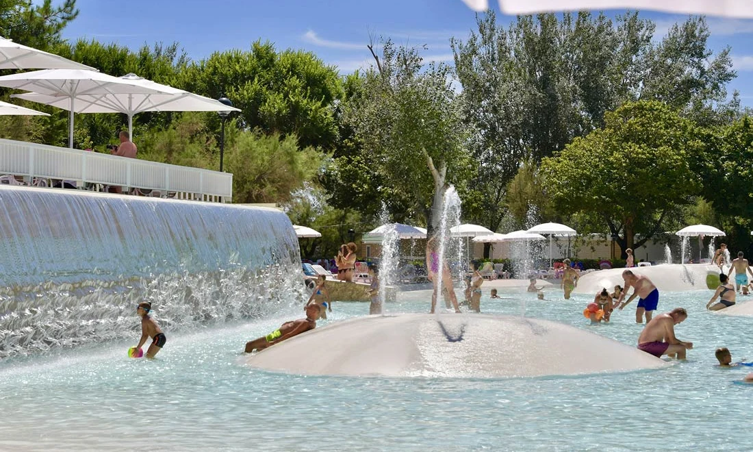 Children playing in shallow pool with fountains and waterfall near shaded lounge area, surrounded by trees at Isamar