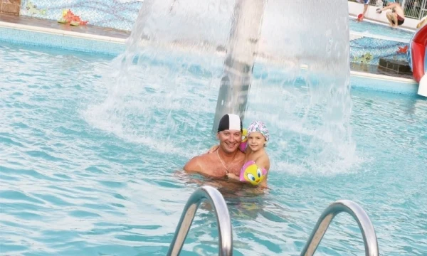 Two people swim under a fountain in a busy pool area, surrounded by other swimmers and pool features at Oasi