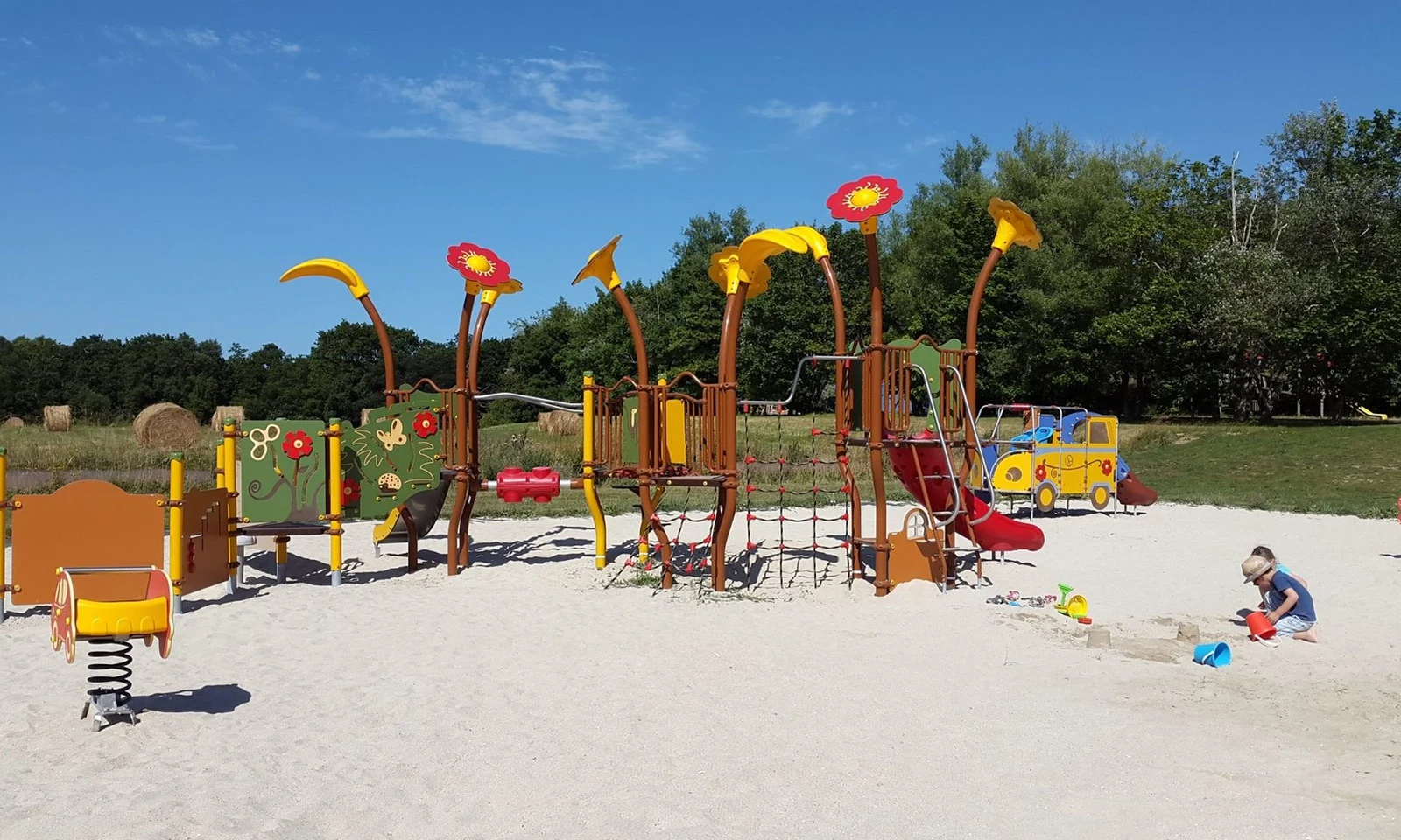 Playground with colorful equipment featuring flower-themed designs; children playing in a sandpit; grassy field with trees and hay bales in the background at Domaine de Mesqueau