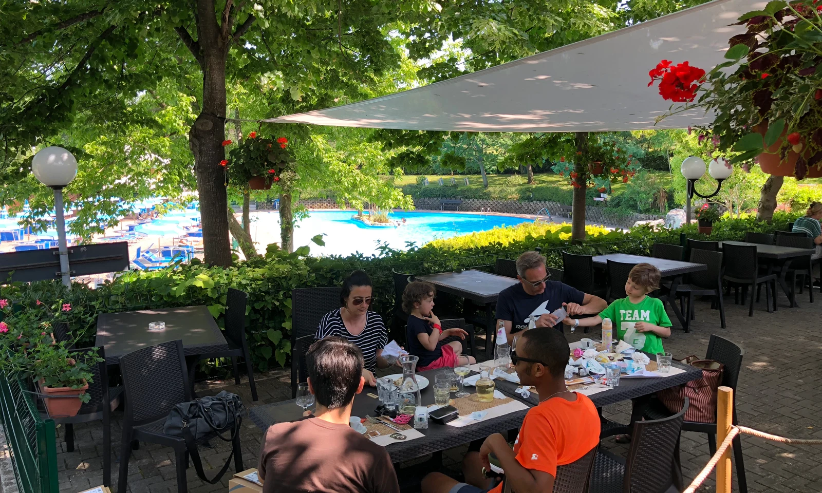 People eating at an outdoor patio surrounded by trees and near a swimming pool at Centro Vacanze San Marino