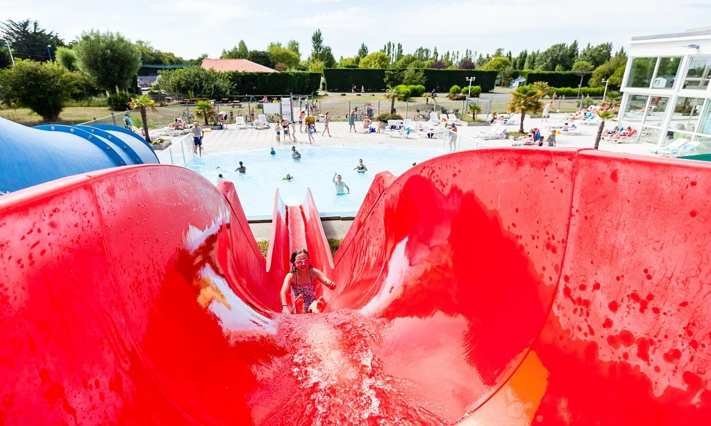 Child slides down a large red waterslide, landing in a crowded pool area with people relaxing and swimming at Les Amiaux