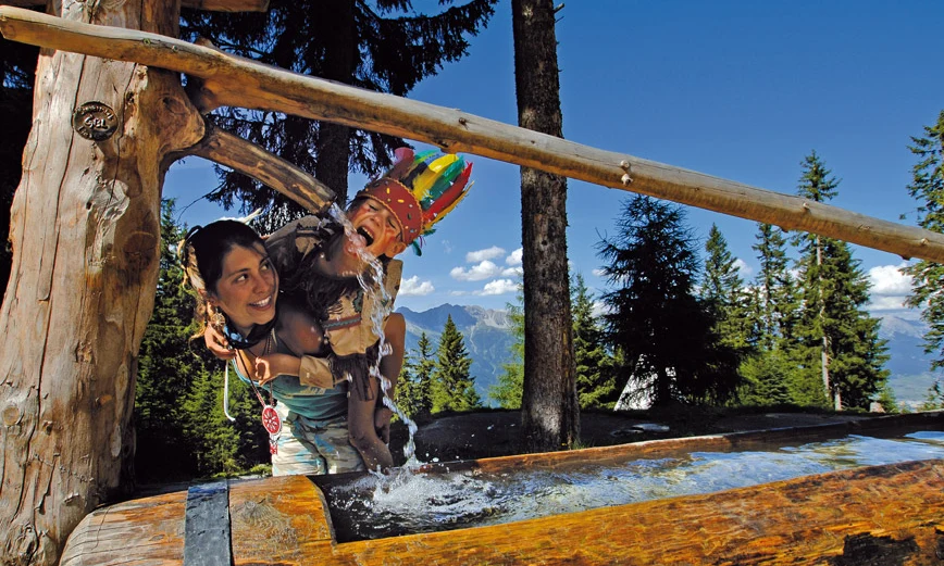 Child wearing colorful feather hat drinks from wooden water trough, surrounded by forested mountain scenery at Natterer See