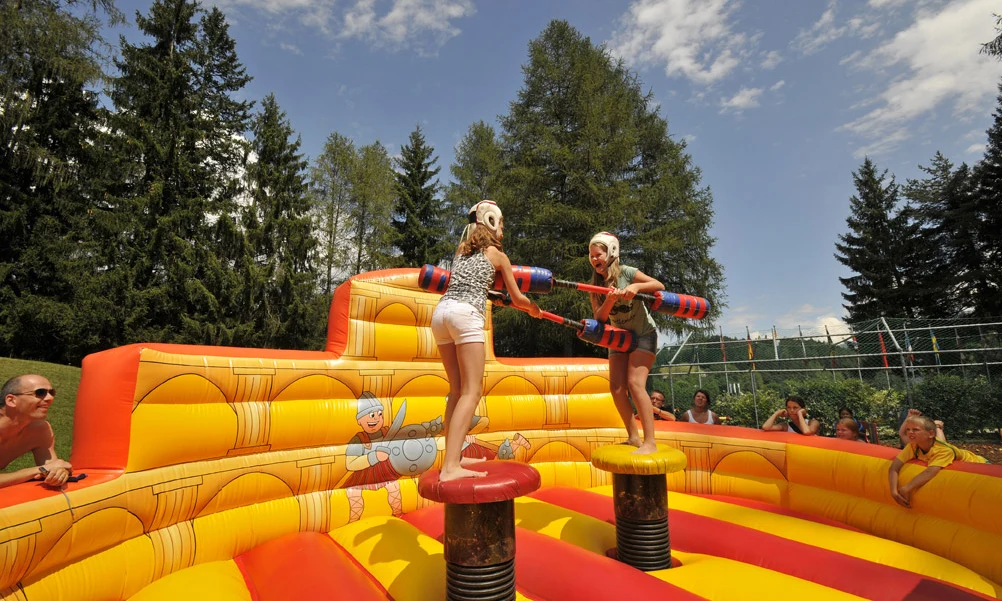 Two people joust with padded sticks on inflatable pedestals, surrounded by an audience and trees at Natterer See