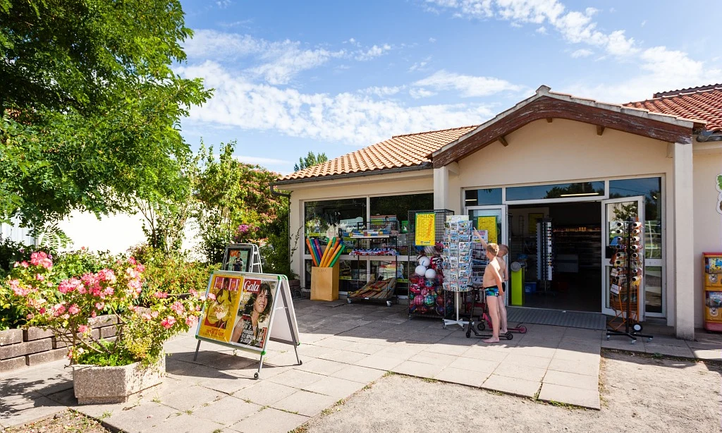 Storefront displays various items; children stand outside it; sunny day with flowers and trees around at Les Amiaux