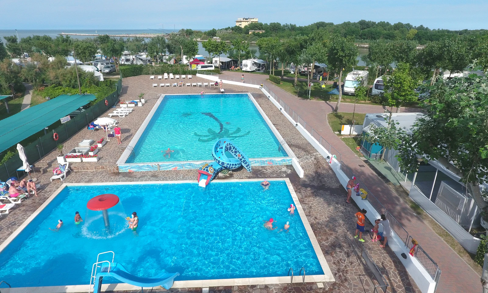 Multiple people swimming and relaxing around two large pools, surrounded by lush greenery and RVs at Oasi