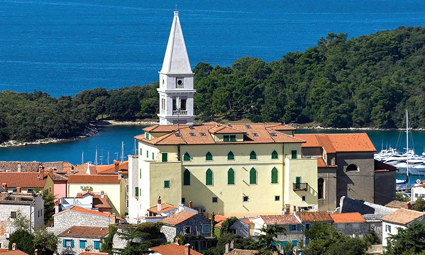 Bell tower rising above a yellow building and terracotta rooftops, with a forest and blue sea in the background at Orsera