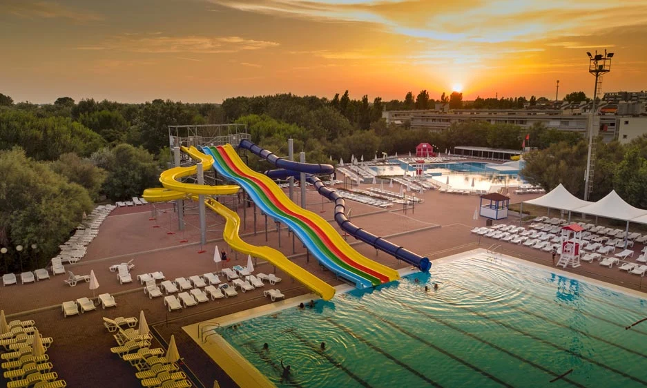 Waterslides lead to a large pool surrounded by lounge chairs and tents, with a sunset sky in the background at Isamar