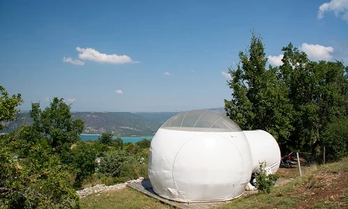 A white, dome-shaped tent situated on a grassy hilltop with trees and a distant lake view at de l'Aigle