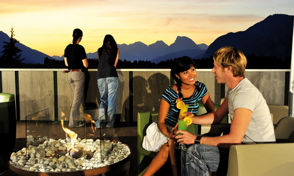 A couple sits by a fire pit with drinks at sunset, mountains in the background at Natterer See