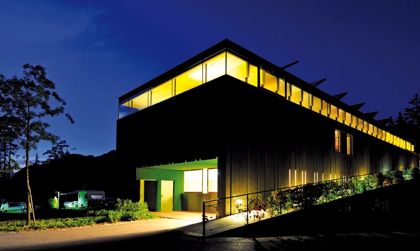 A modern, illuminated building stands amidst trees with camping trailers nearby at Natterer See