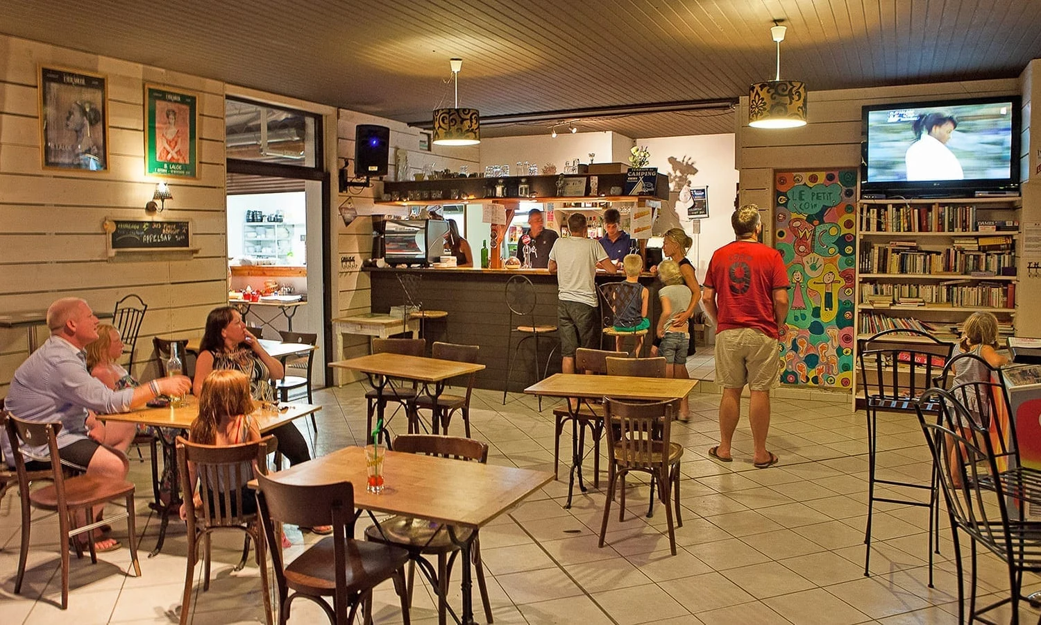 People relaxing and socializing in a cozy cafe, with some watching TV and others ordering at the counter at Ardeche Camping 