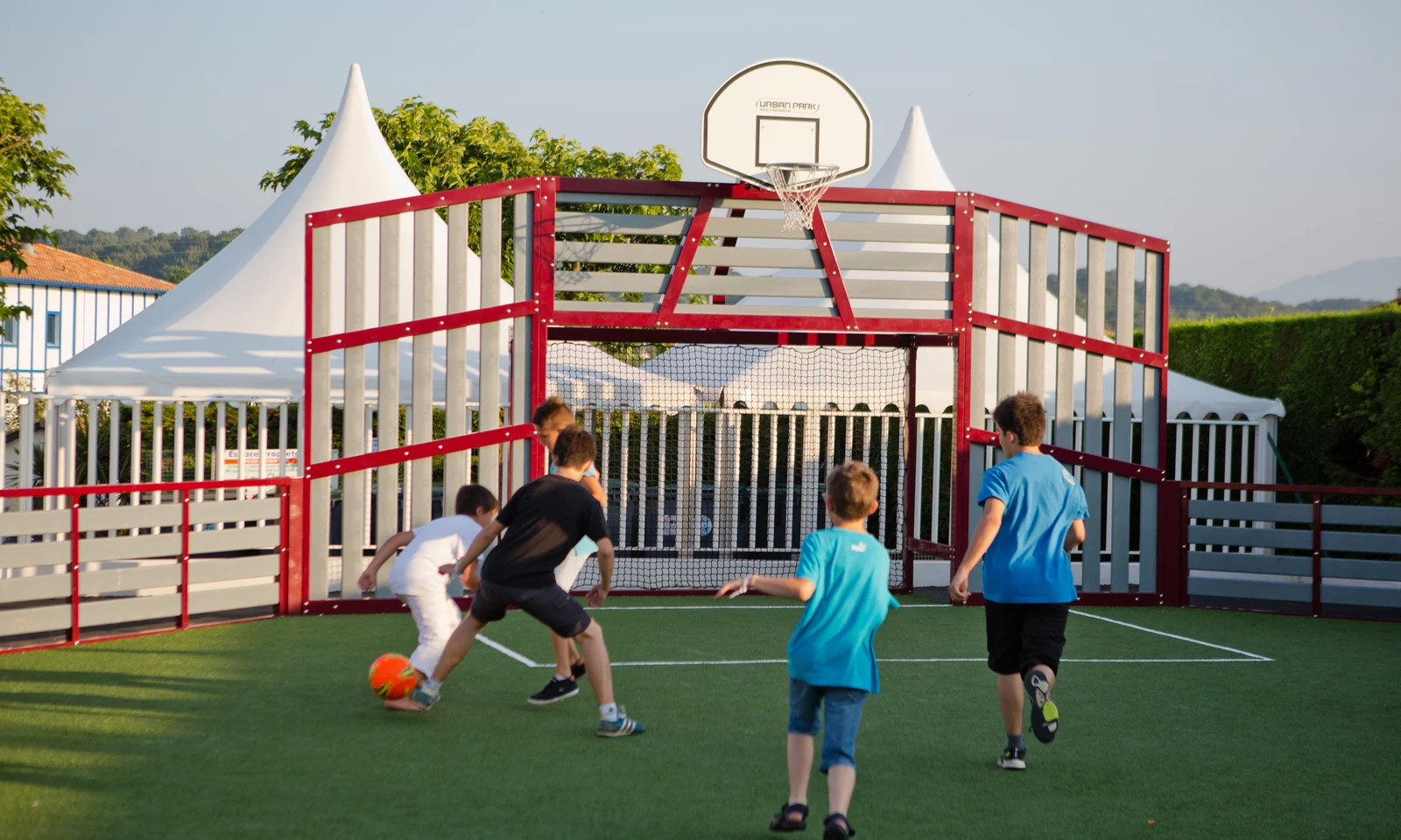 Children play soccer on an artificial grass court, surrounded by white tents and trees at Ur-Onéa