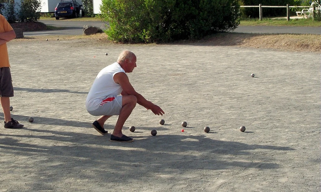 A man playing pétanque, bending down to throw a ball, on a sandy playing field with a parked car and house nearby at Les Amiaux