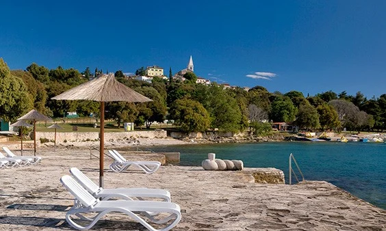 White lounge chairs are arranged under straw umbrellas on a stone platform overlooking a forested coastal town at Orsera