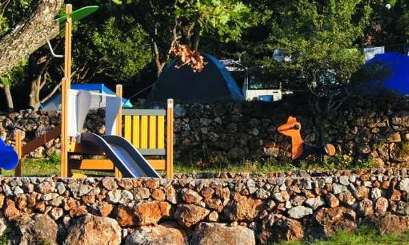 Children's playground with slide, climbing net, and seesaw in front of stone wall and tents at de l'Aigle