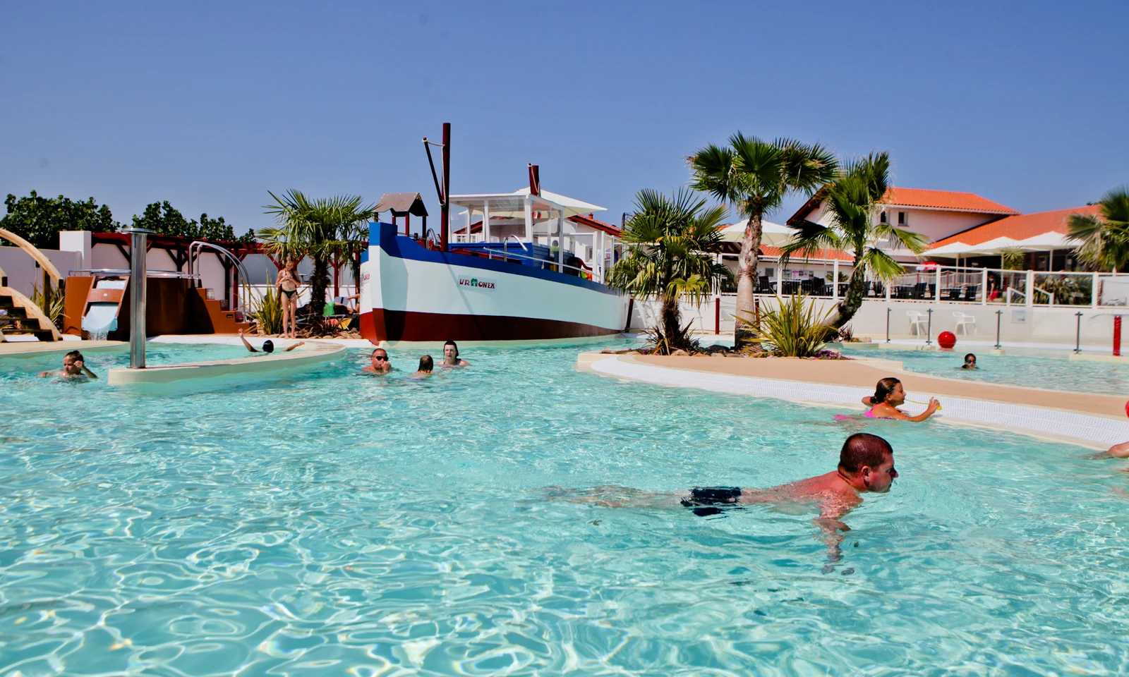 People swim in a clear pool with a large boat structure and palm trees nearby at Ur-Onéa