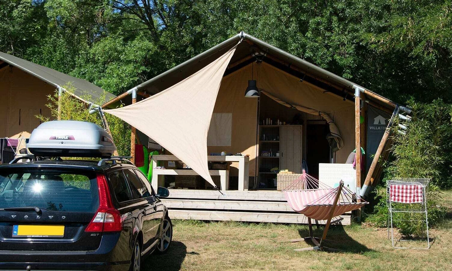 Tent with attached awning sheltering patio and furniture, placed in a forested campsite with a parked car and hammock at Ardeche Camping 