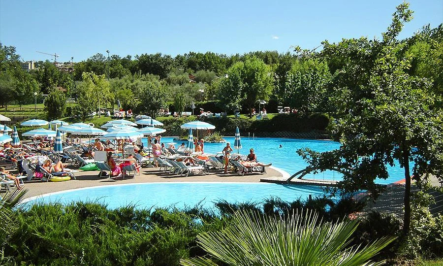 People relaxing on lounge chairs and swimming in a large pool surrounded by trees and umbrellas at Centro Vacanze San Marino