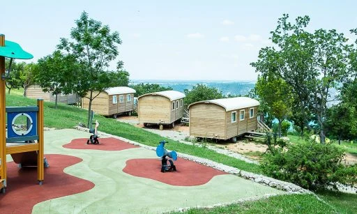 Wooden play structure with climbing feature and slide situated near rustic cabins amidst trees and greenery at de l'Aigle