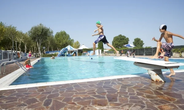 Two children jump off a diving board into a busy outdoor swimming pool with people and greenery around at Oasi