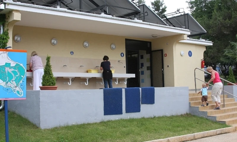 Women washing dishes at an outdoor sink; another woman helps a toddler on stairs leading to a restroom at Orsera