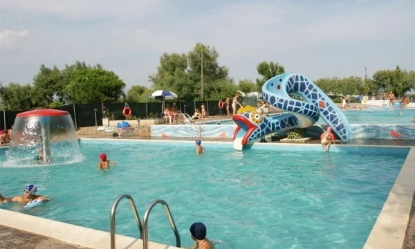 Children swim and play around colorful water slides in a pool area surrounded by trees and distant people at Oasi