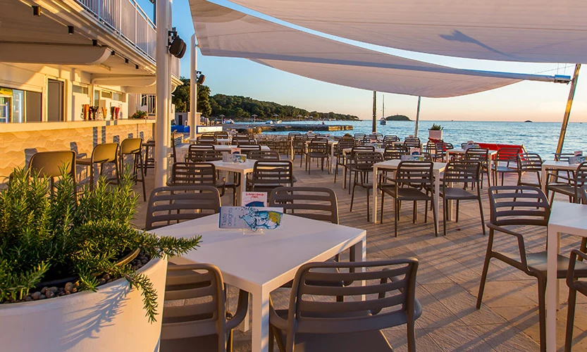 Tables and chairs arranged under canopies by a seaside cafe, with a planter and sunset view at Orsera