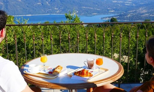 Two people sit at a table enjoying breakfast, overlooking a scenic lake and mountainous landscape at de l'Aigle