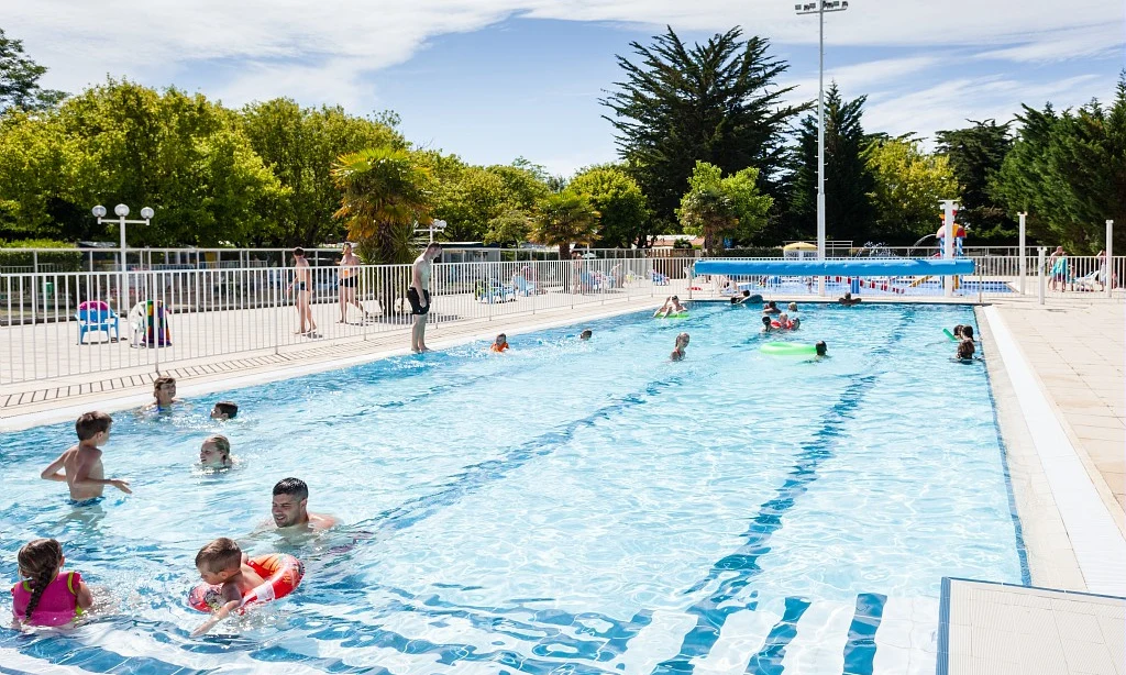 People swimming and playing in a bright blue pool surrounded by trees and a fence at Les Amiaux