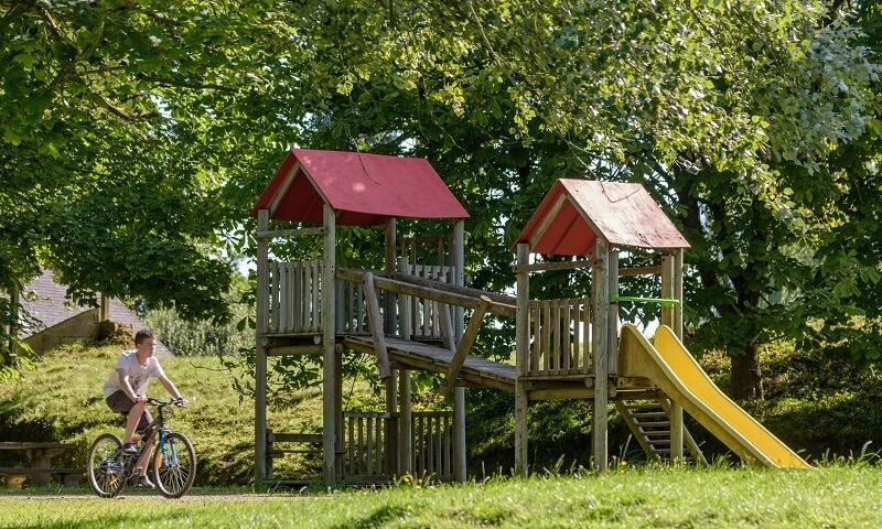 Wooden playground structure with a slide, cyclist passing by, lush greenery and trees surrounding at Domaine de Mesqueau