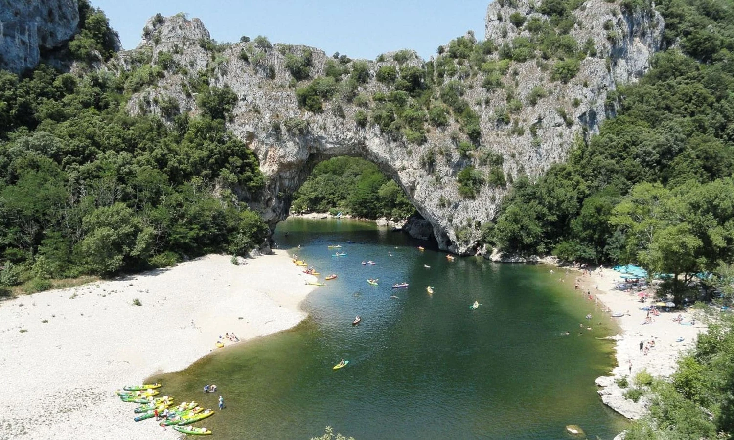 Kayaks are floating on a river surrounded by rocky cliffs and wooded areas, with a natural stone arch overhead at Ardeche Camping 