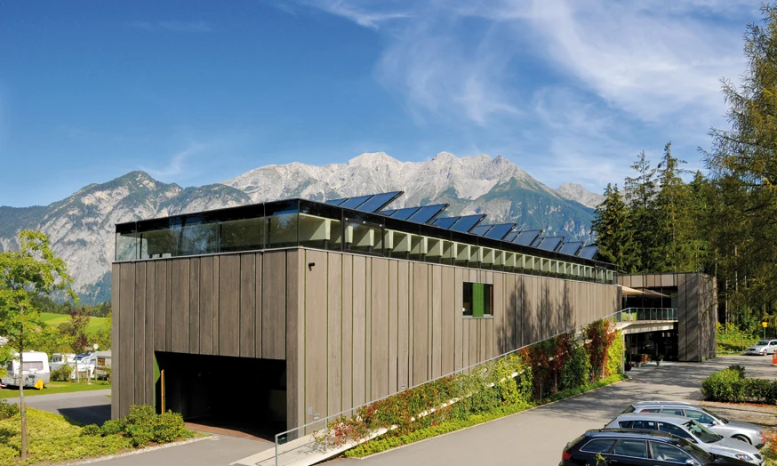 Modern building with solar panels sits by parked cars and lush mountains in scenic natural setting at Natterer See