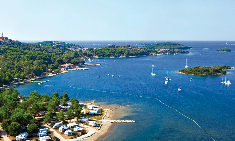 Campsite by a forested coastline with several caravans parked along the shore, sailboats anchored offshore at Orsera