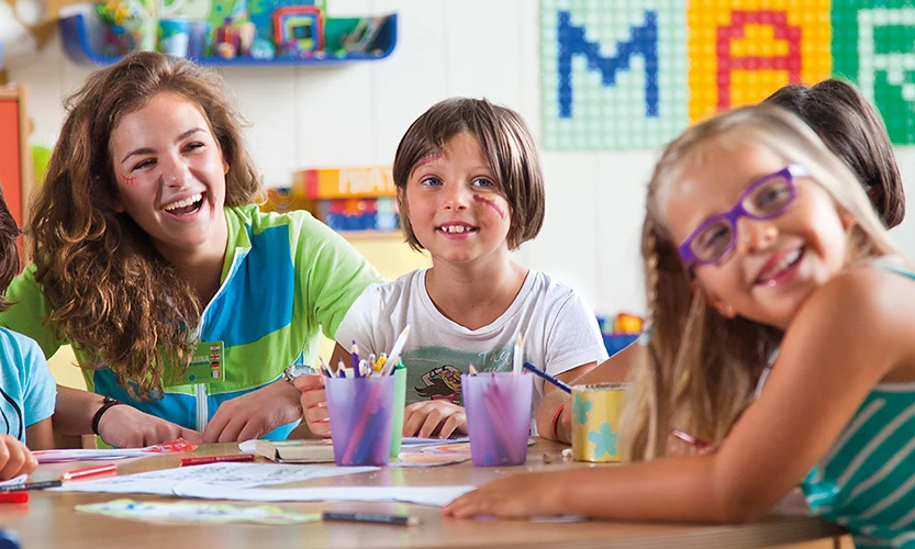 Children and an adult are smiling and drawing together at a colorful indoor arts and crafts table at Orsera