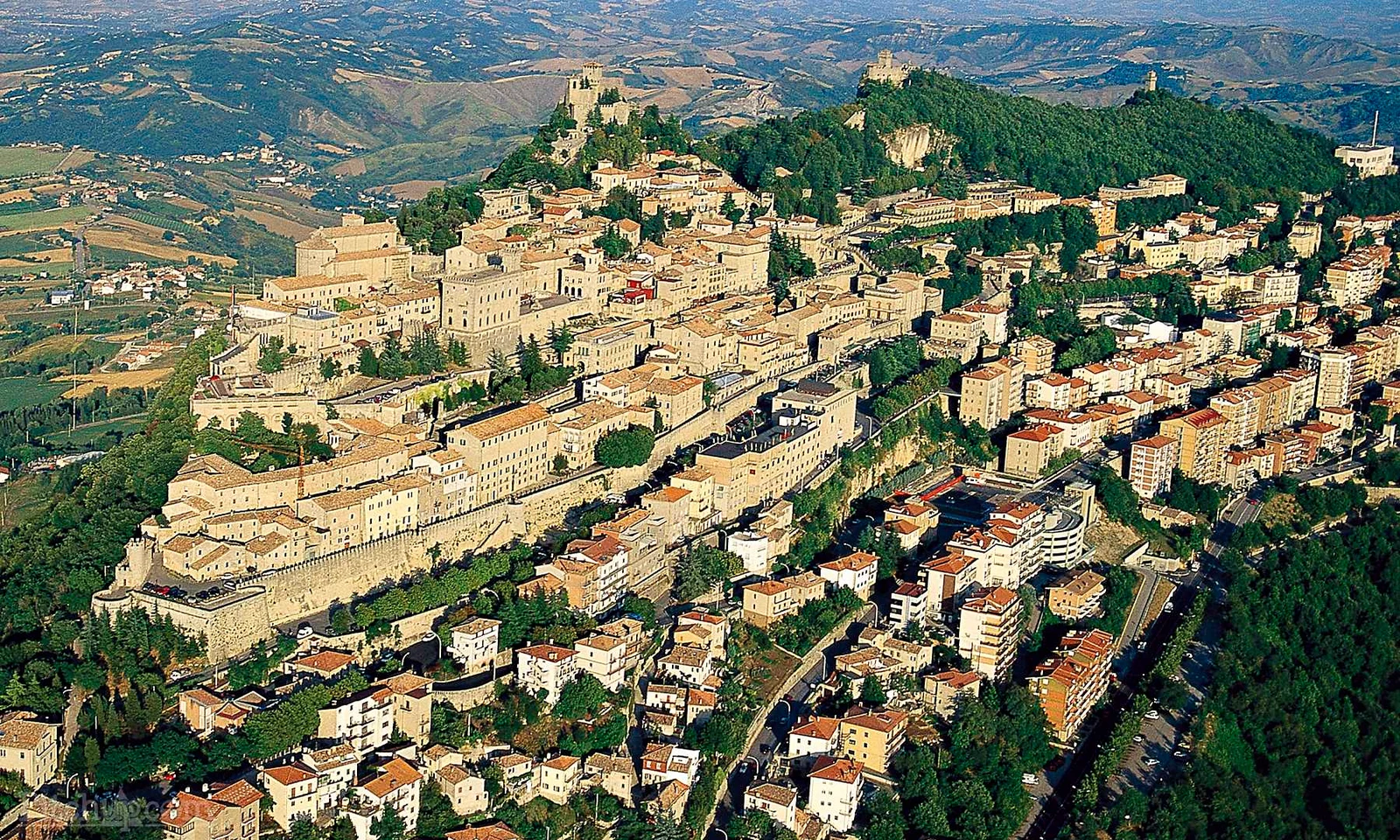 Aerial view of a hillside town with historic buildings and lush greenery, surrounded by vast, rolling landscapes at Centro Vacanze San Marino