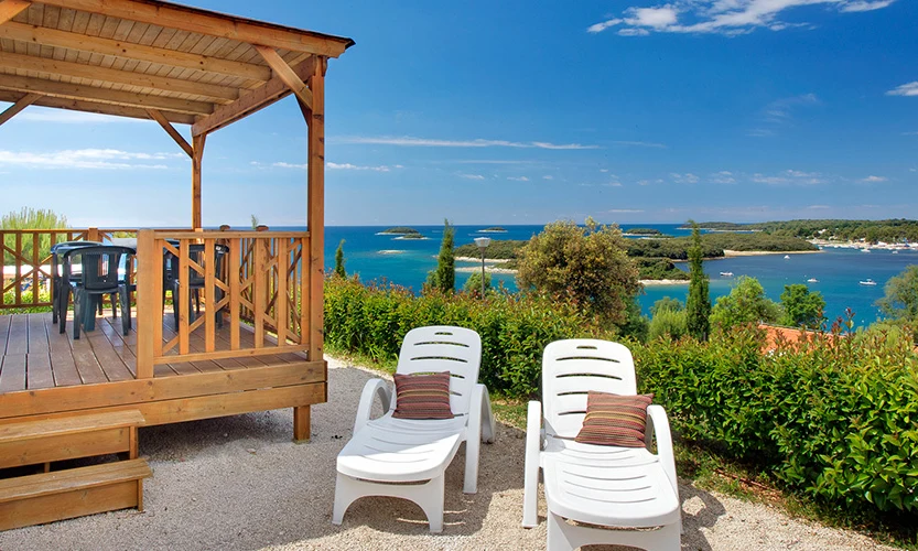 Wooden veranda with plastic chairs near two sun loungers facing a scenic coastline view with green shrubs and blue water at Orsera