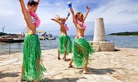 Three women wearing hula skirts and leis dancing enthusiastically by the seaside on a sunny day at Orsera