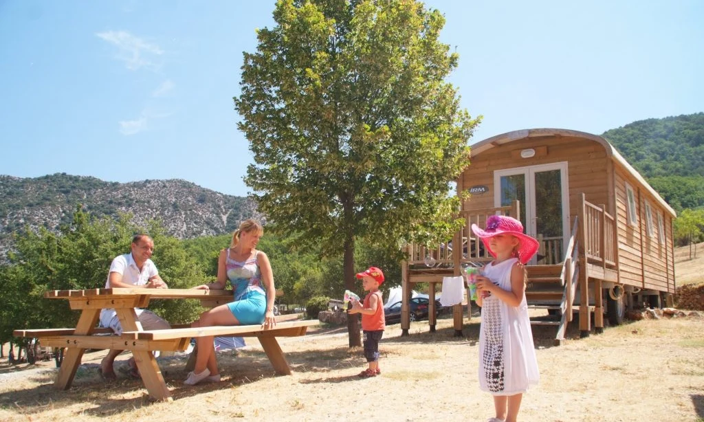 Family relaxing and playing near a wooden cabin with mountains in the background at de l'Aigle