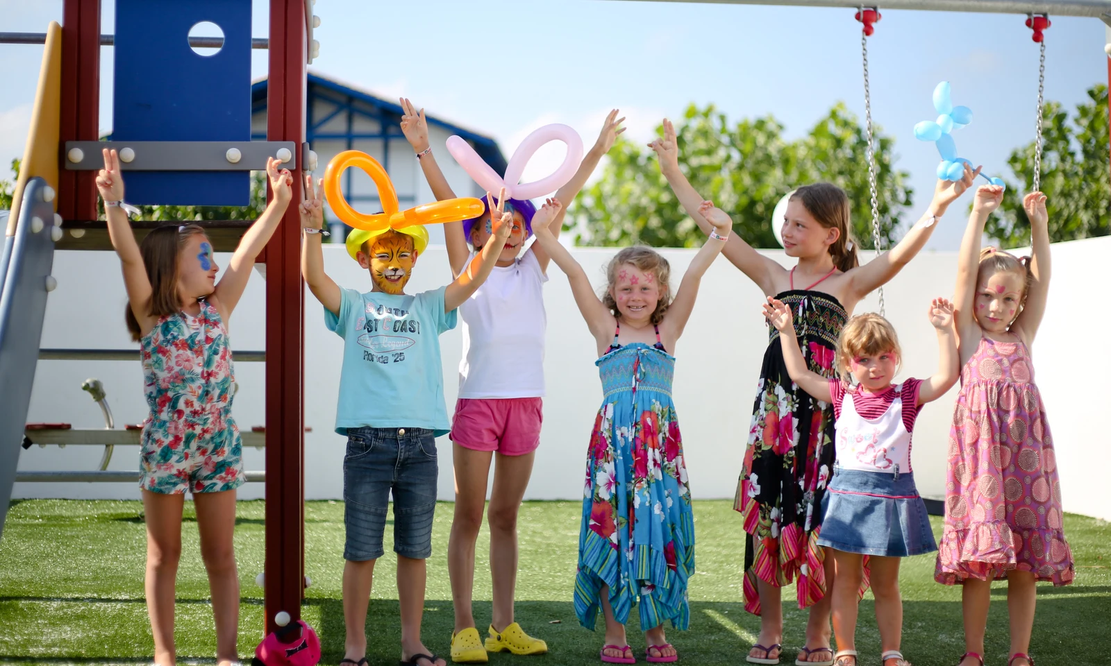 Children with face paint and balloon hats, raising arms happily on a playground with green grass and trees at Ur-Onéa