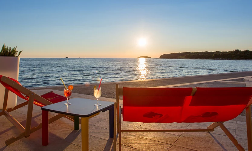 Two red deck chairs face the ocean at sunset, with cocktails on a small table at Orsera