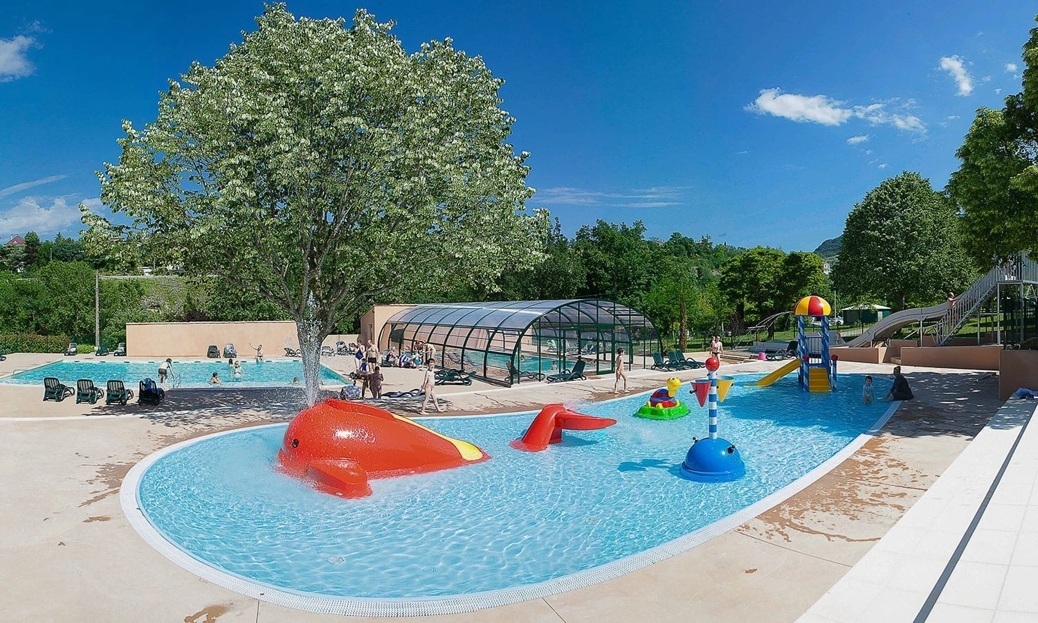 Children playing in a pool with water slides and toys surrounded by trees and lounge chairs at Ardeche Camping 