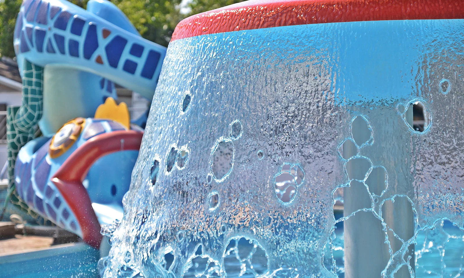 Fountain spraying water, with colorful inflatable climbing structure in the background, surrounded by trees at Oasi