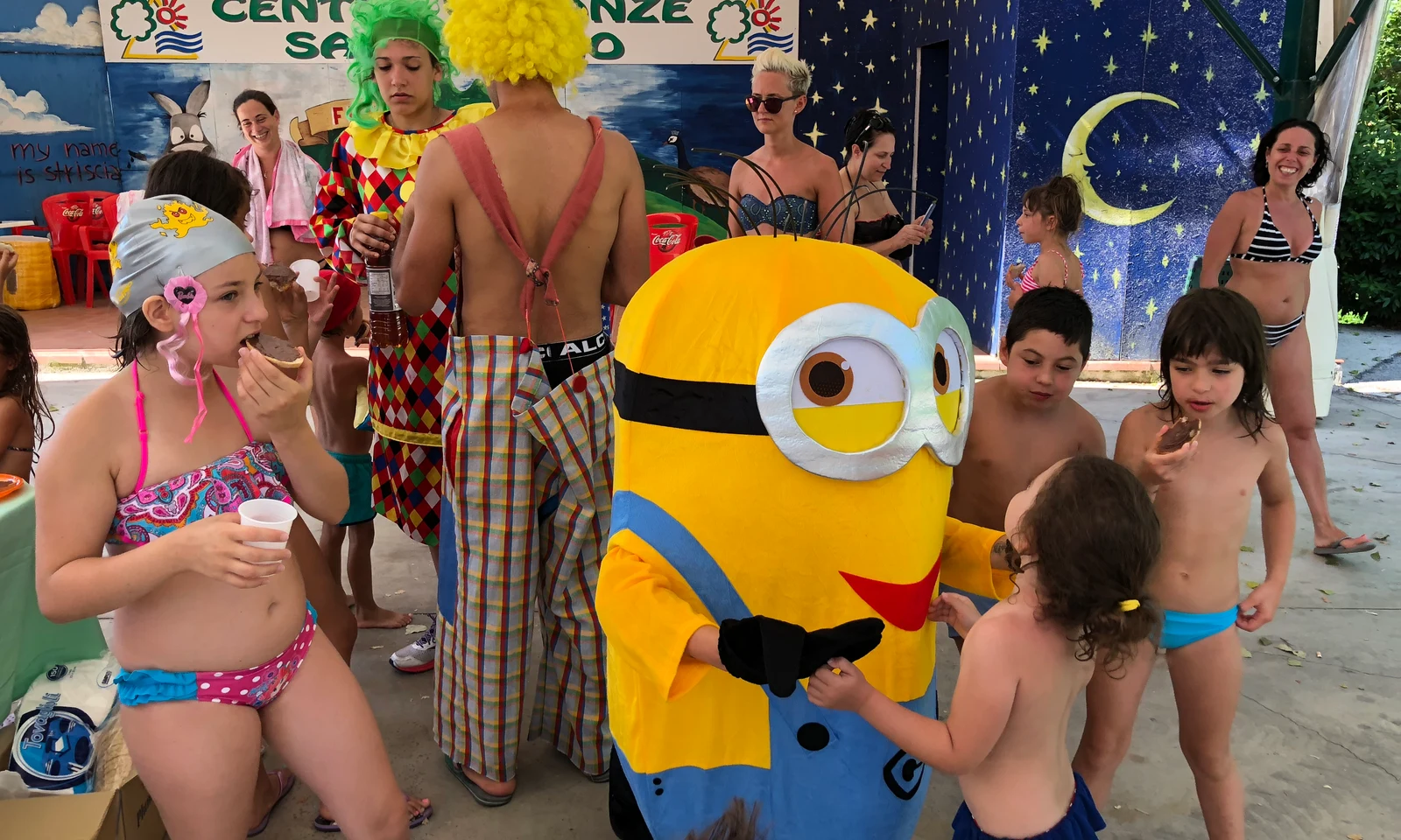 Minion mascot greeting children and a clown eating snacks in a colorful outdoor area with a starry night mural at Centro Vacanze San Marino