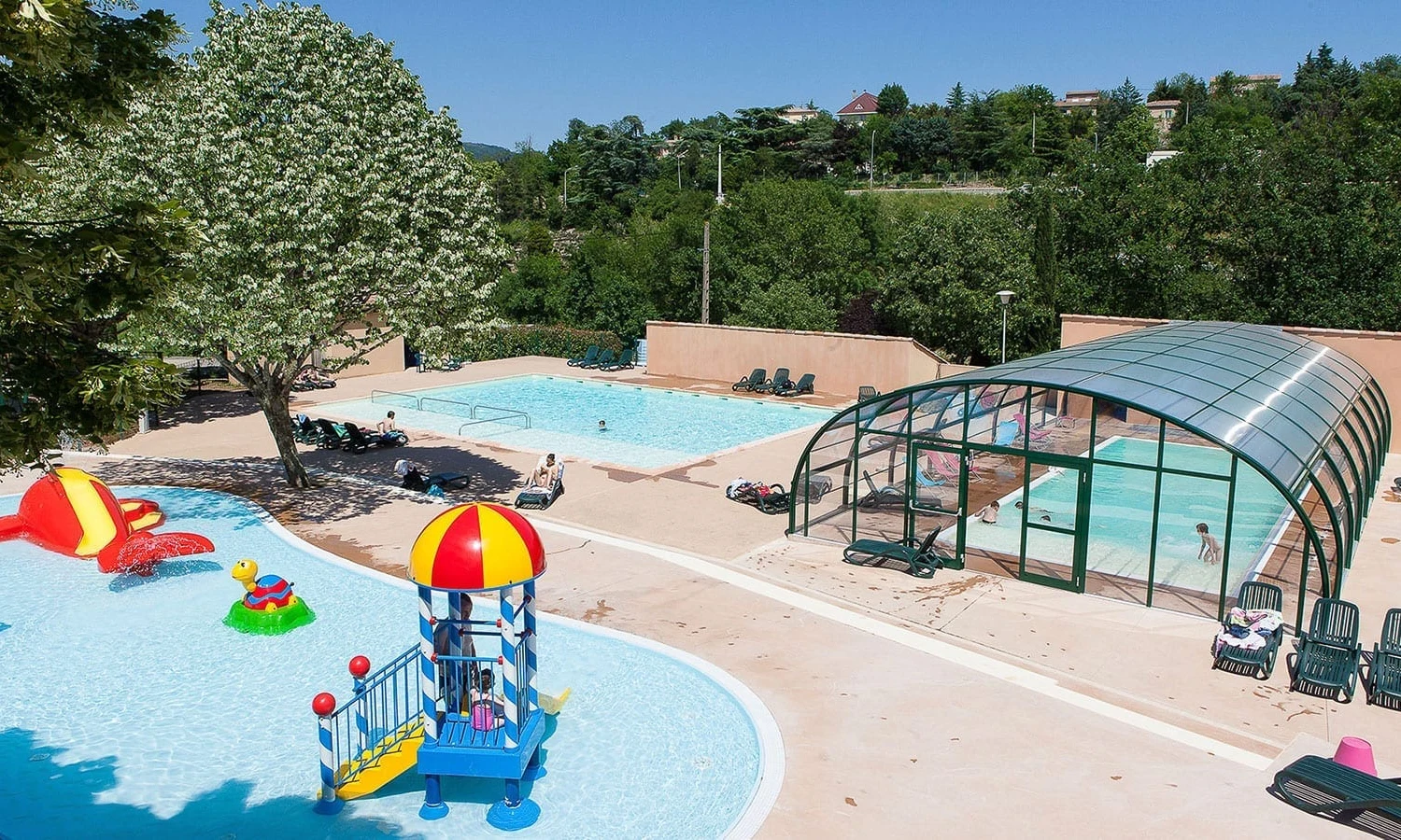 Swimming pools with slides, children playing; large tree nearby; loungers scattered around; semi-enclosed pool structure; forested background at Ardeche Camping 