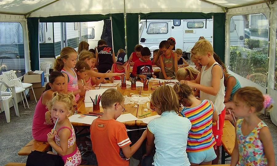 Children drawing and coloring on a table under a canopy with caravans visible outside at Leï Suves