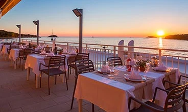 White-clothed tables arranged for dinner overlooking a tranquil sea at sunset on a restaurant patio at Orsera