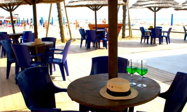 Table with two green drinks and a hat on it, surrounded by chairs, under thatched umbrellas on a sandy beach at Oasi